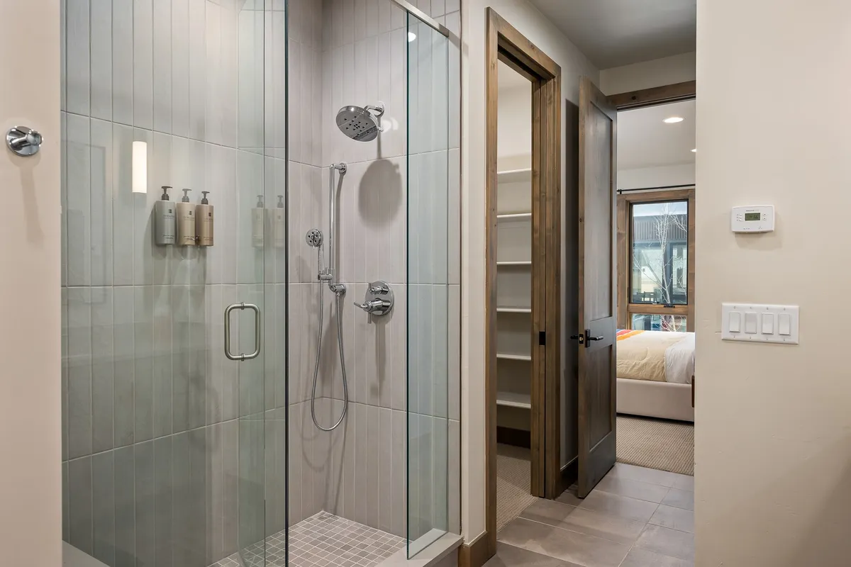 A modern bathroom with a glass shower enclosure, warm wood accents, and sleek fixtures. The open door provides a glimpse into the adjacent bedroom, creating a seamless, restful suite. Natural light highlights the bathroom's inviting, spa-inspired feel.