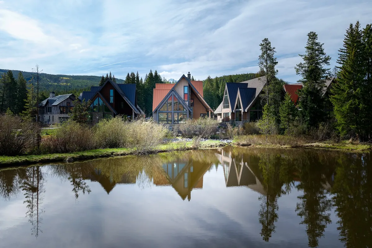 A tranquil exterior scene with modern architecture reflecting on calm water, surrounded by lush trees. The scene suggests a place for walking, nature photography, or simply enjoying the changing seasons by the water.