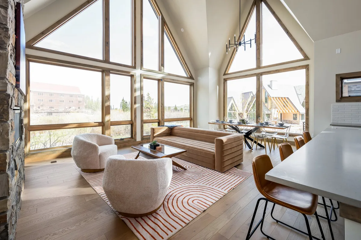 A contemporary great room featuring a modern sofa, textured armchairs, a dining table, a stylish chandelier, and large windows. The room is bright and airy, perfect for relaxing, socializing, dining, and enjoying the natural views.