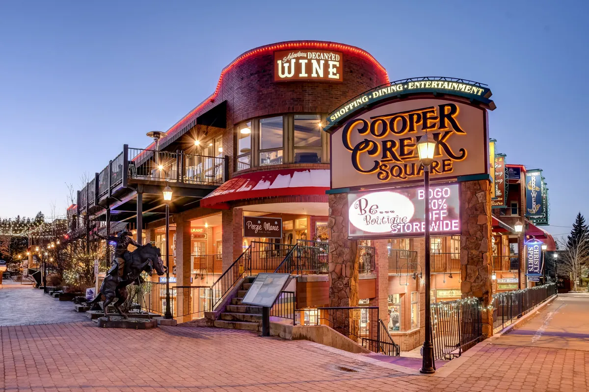 A festive downtown scene featuring the Cooper Creek Square sign, outdoor stairs, and vibrant wine signage amidst a backdrop of shops and eateries—illuminated with lively colors and bustling activity.