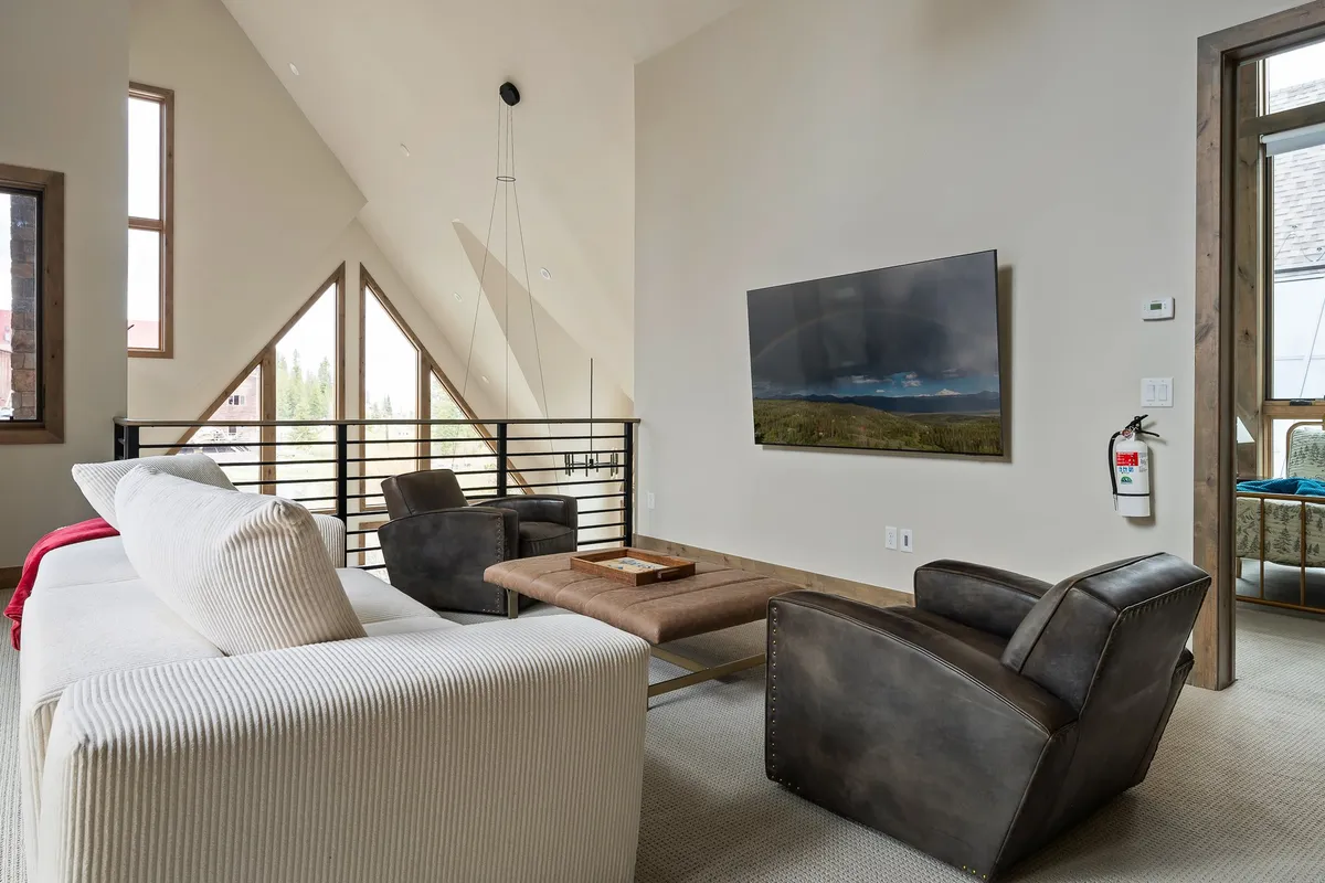 This image captures a modern living space with large geometric windows, a contemporary coffee table, and a sizable television. Sunlight pours in, creating an inviting, cozy spot ideal for relaxing after a day outdoors or hosting movie nights. The windows suggest a strong connection to the surrounding scenery, echoing the theme of retreating to tranquil views.