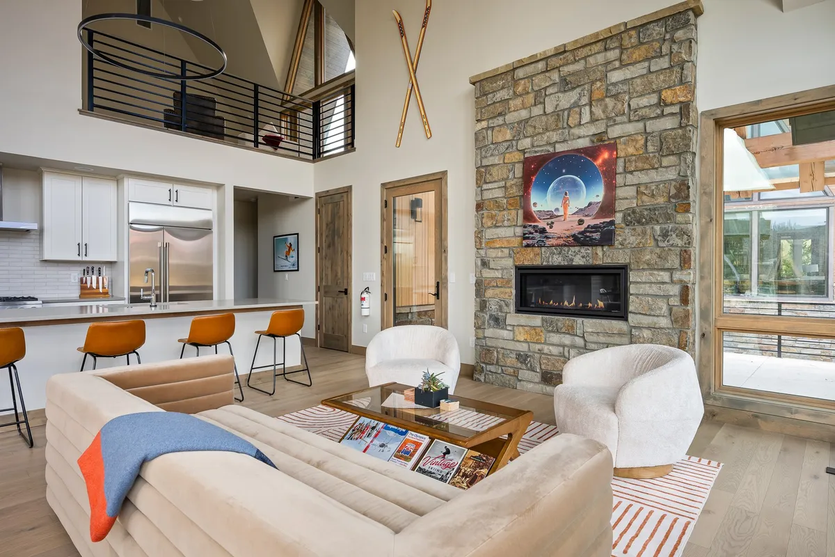 The image highlights a spacious open-concept living area with a striking custom stone fireplace as the centerpiece. A modern couch faces the fireplace, with bar stools at the kitchen island visible in the background, suggesting a seamless flow between the living room and kitchen. Artwork above the fireplace and abundant natural light create an inviting, stylish atmosphere fit for social gatherings.