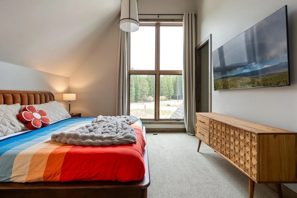 A comfortable bed with colorful bedding is positioned beside a large window that frames a lush, forested view. The room also features a TV stand and hints of warmth from natural wood accents, suggesting a restful retreat with a seamless connection to nature.