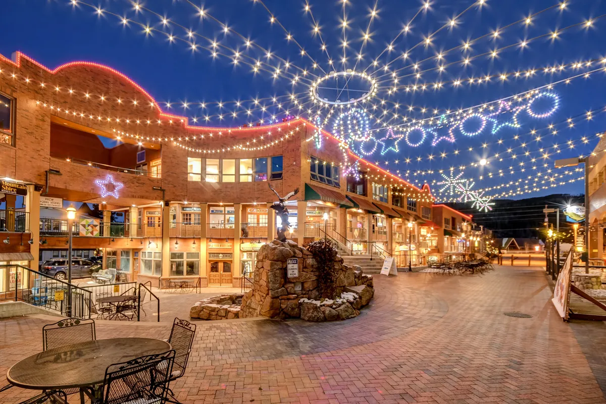 A lively downtown plaza illuminated by overhead string lights and decorative stars, with a central rock fountain and inviting outdoor seating. The scene buzzes with people dining, shopping, and socializing, evoking a festive, welcoming urban vibe both day and night.