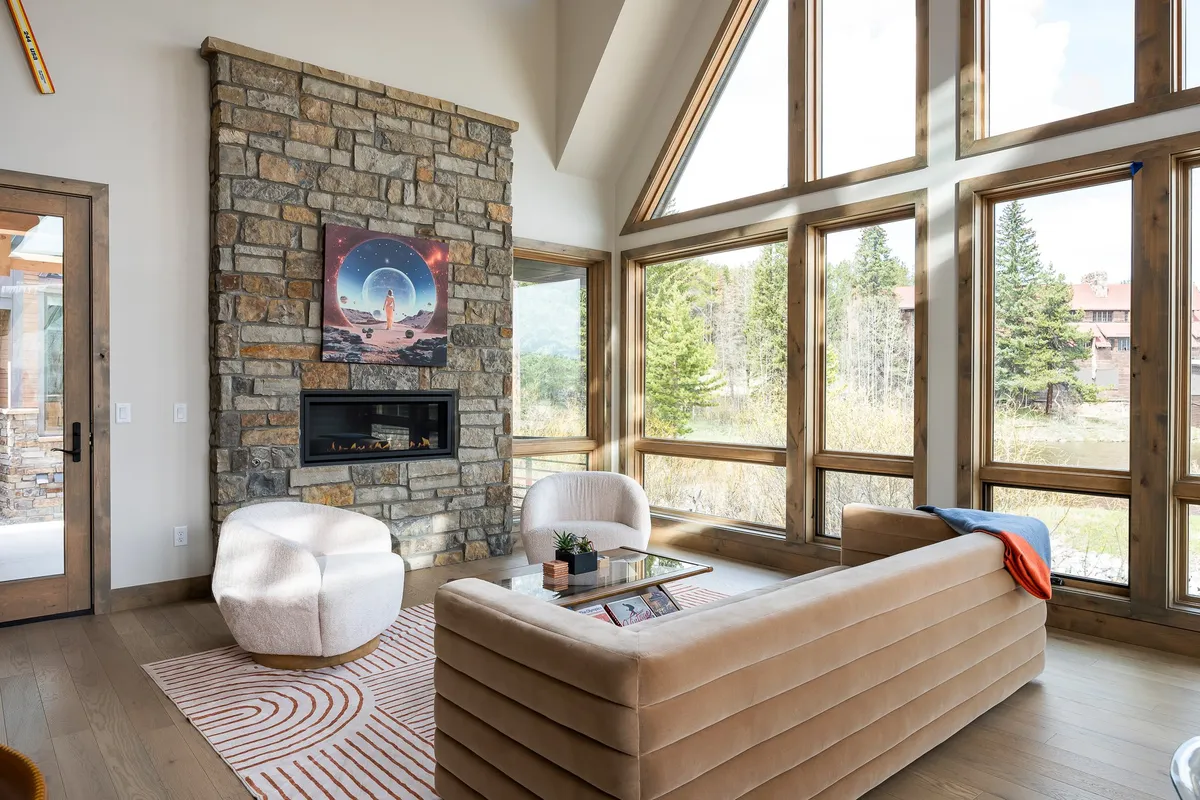 A spacious living area with a stunning stone fireplace as its centerpiece, flanked by large windows that fill the room with natural light. Modern furniture and unique artwork create a sophisticated yet warm ambiance, making it ideal for relaxation or social gatherings after a day on the mountain.