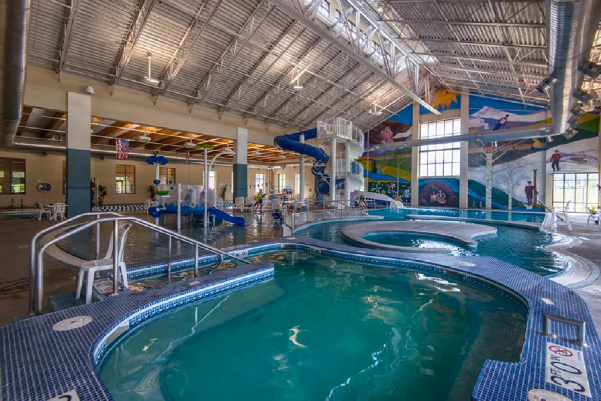 An energetic indoor pool scene with children and families enjoying water slides, a large swimming pool, vibrant murals, and a designated play area—evoking the atmosphere of a lively rec center.